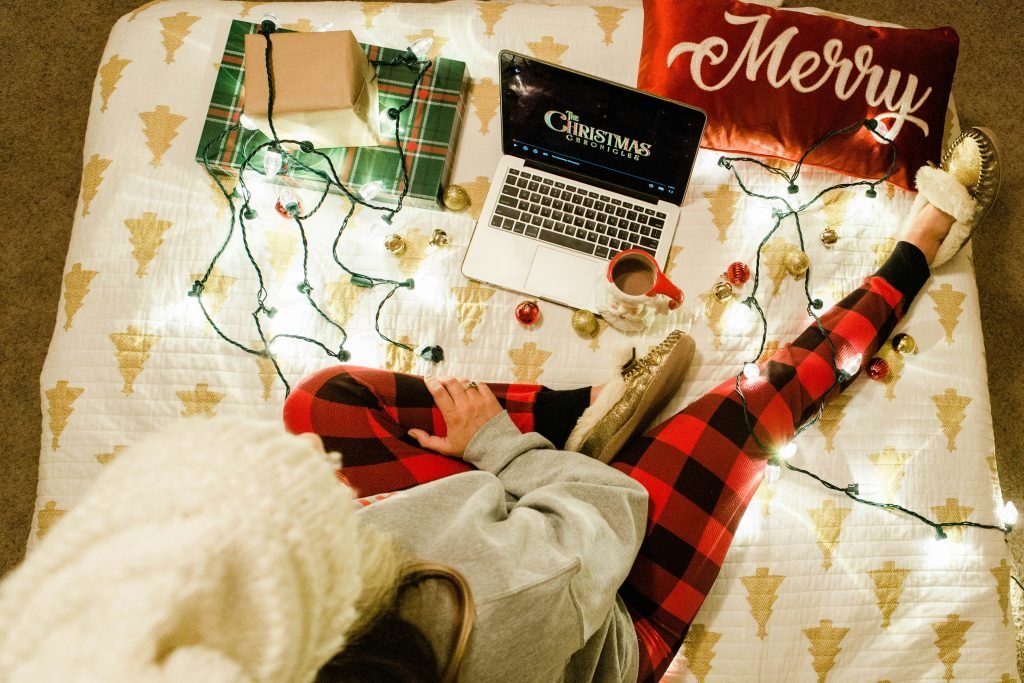 Overhead view of a cozy Christmas scene: a person in red plaid pants with fluffy slippers sits on a bed, string lights, gifts, a mug, and a laptop open to The Christmas Chronicles.