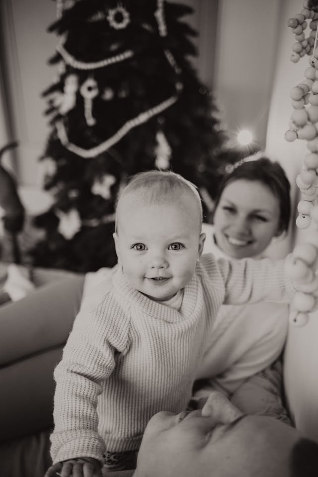 Baby smiling, reaching for a bead garland while a woman watches in a living room with a Christmas tree behind them (black and white).