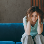 Young woman sits on a blue couch with her head in her hands, looking distressed or tired indoors.
