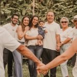 Family and friends form a circle outdoors, holding hands for a group photo with a baby at center.