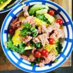 Blue-and-white striped bowl of salad with corn kernels, cherry tomatoes, mixed greens, and flaked tuna on a wooden table.